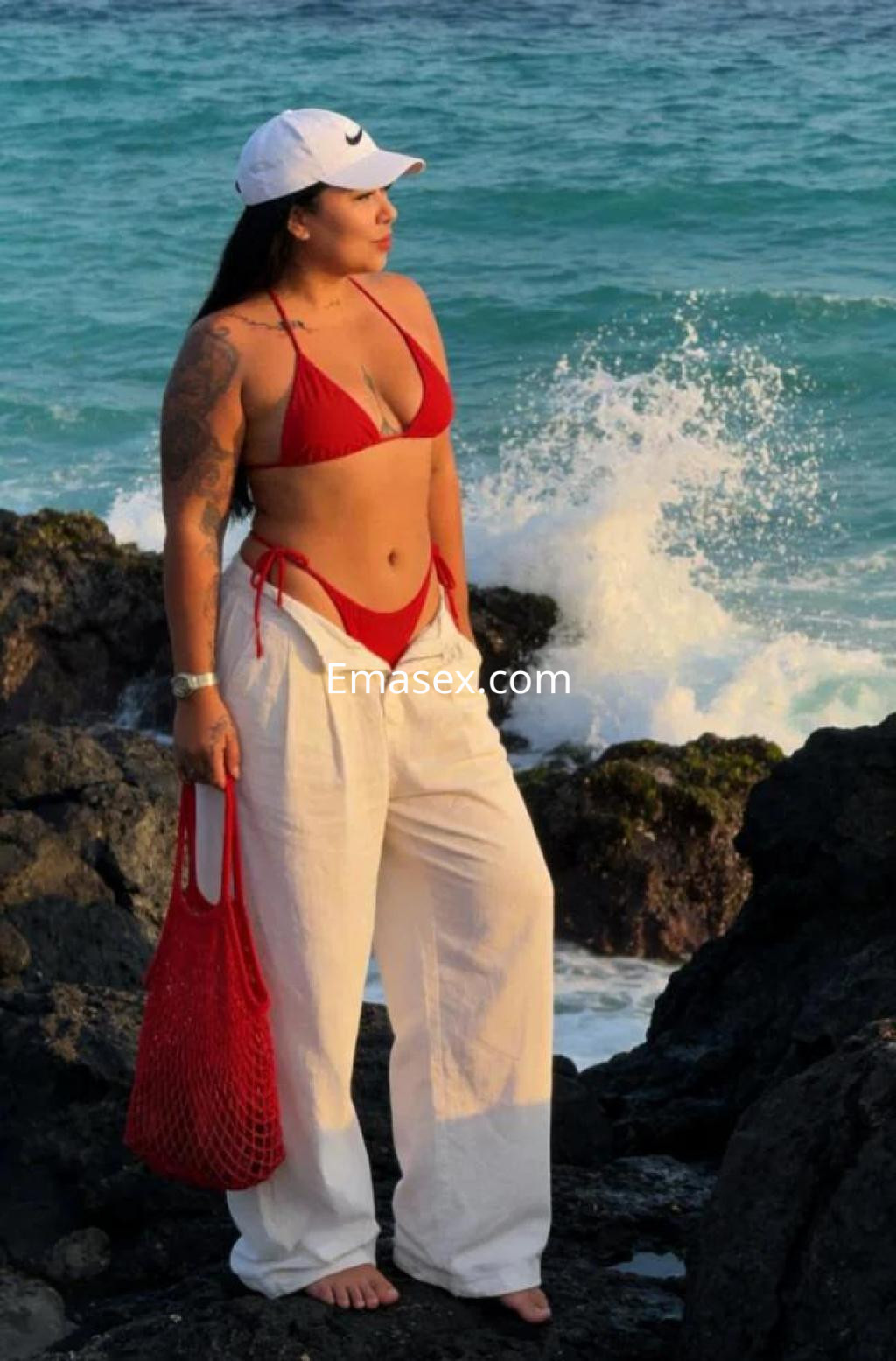 Escort en Las Palmas, con bikini rojo y pantalones blancos, posando frente al mar en una playa rocosa.