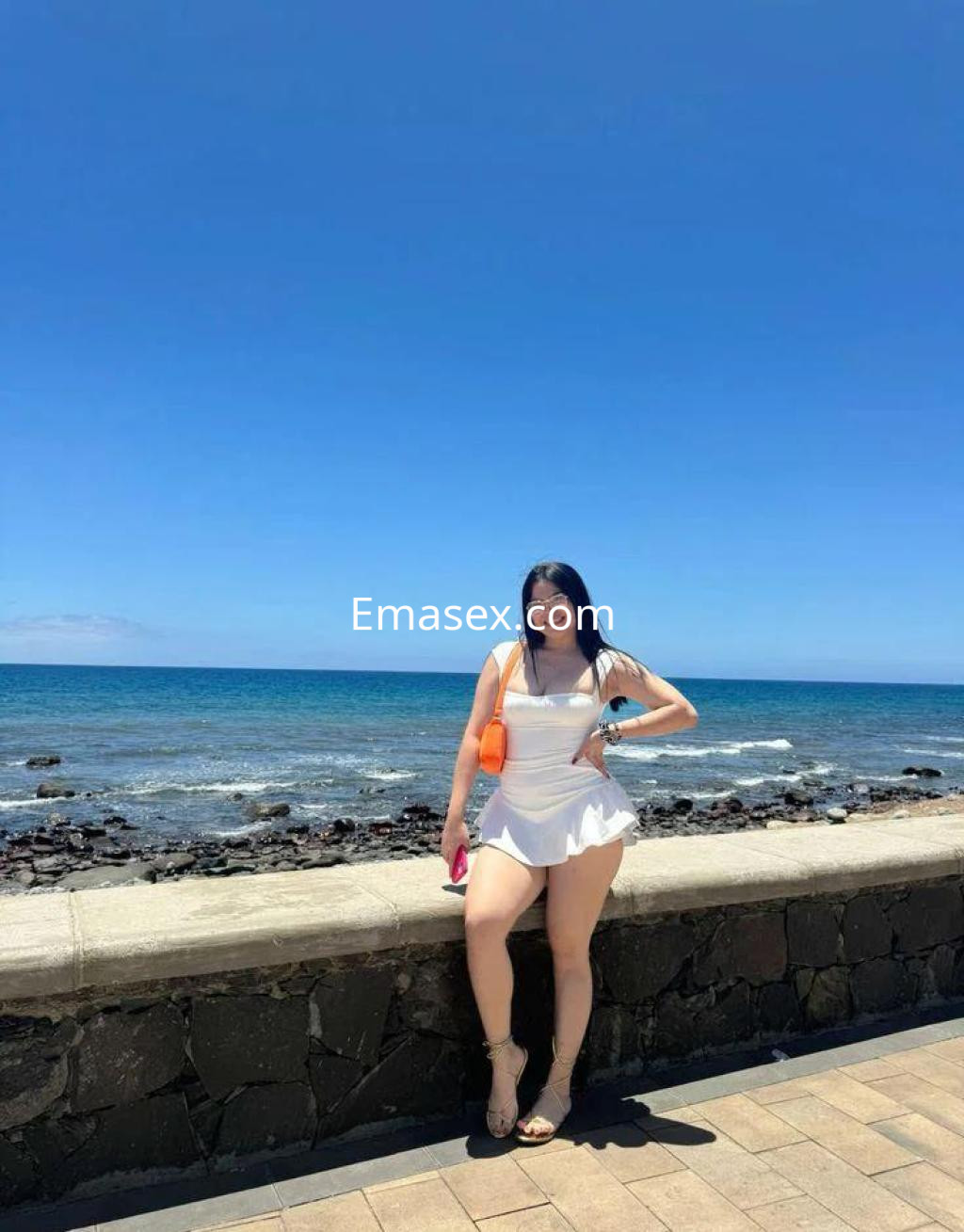 Puta en Tenerife, luciendo un vestido blanco mientras posa junto al mar bajo un cielo despejado.