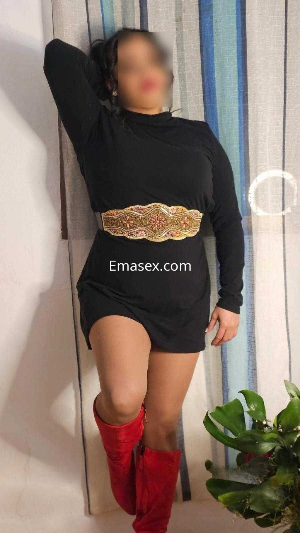 Puta posando en Sevilla con un vestido negro ajustado y botas rojas, apoyada en una pared decorativa.