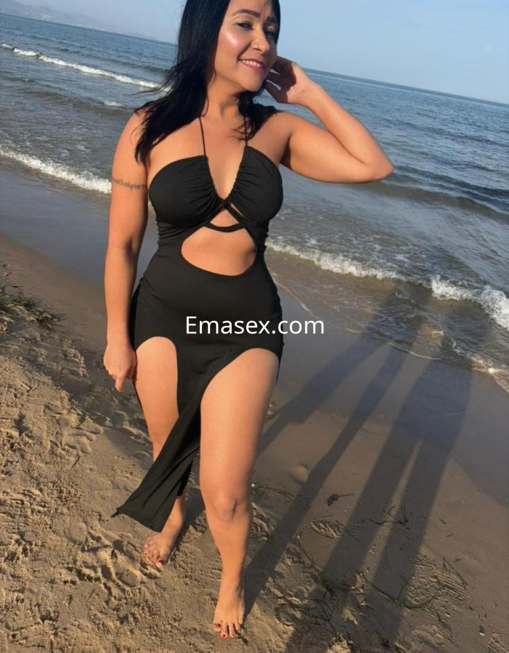 Puta en Toledo con un traje de baño negro, sonriendo en la playa y disfrutando del sol junto al mar.