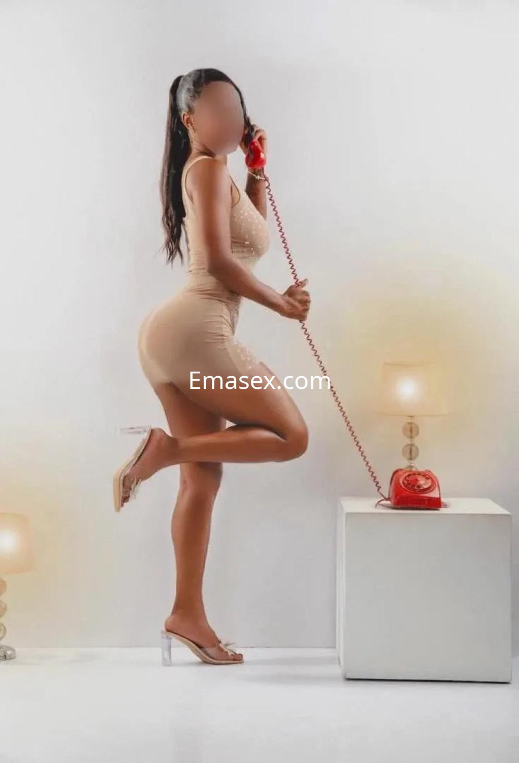 Escort en Valencia, posando con un teléfono retro, viste un ajustado vestido nude y tacones transparentes.