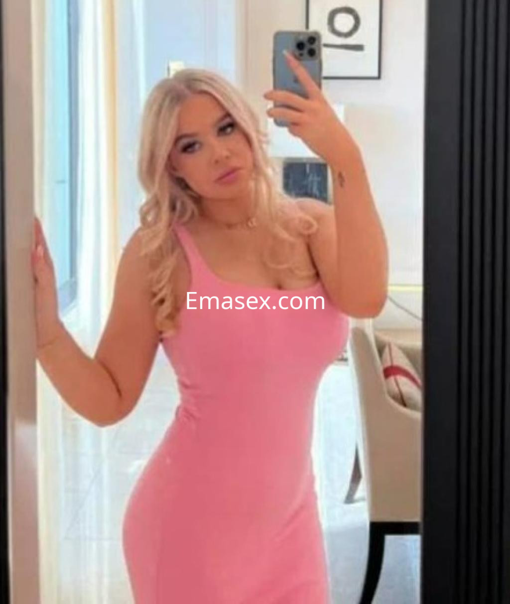 Escort en Barcelona posando frente a un espejo, vistiendo un vestido rosa ajustado y con el cabello rubio ondulado.