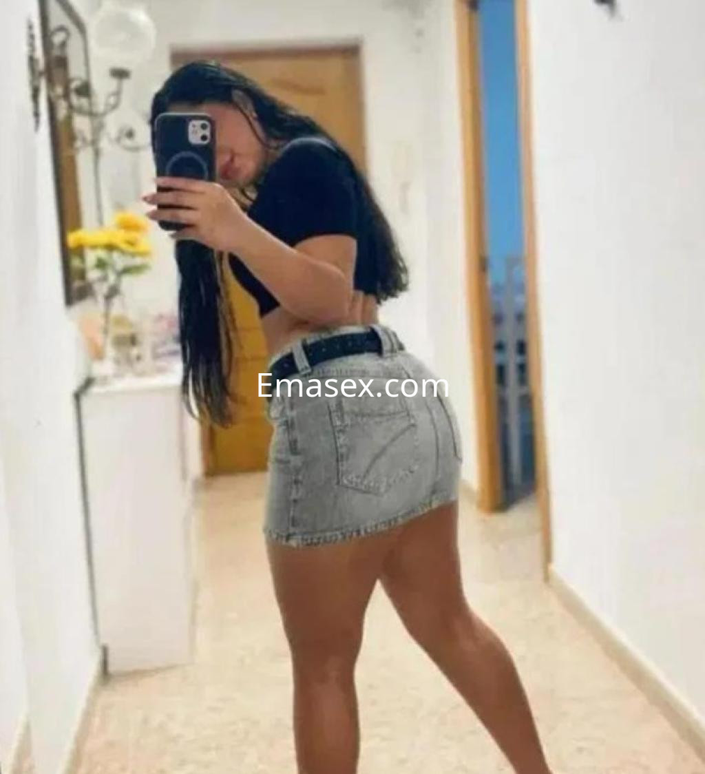 Puta en Burgos posando en un pasillo con una camiseta negra y una falda de mezclilla ajustada.
