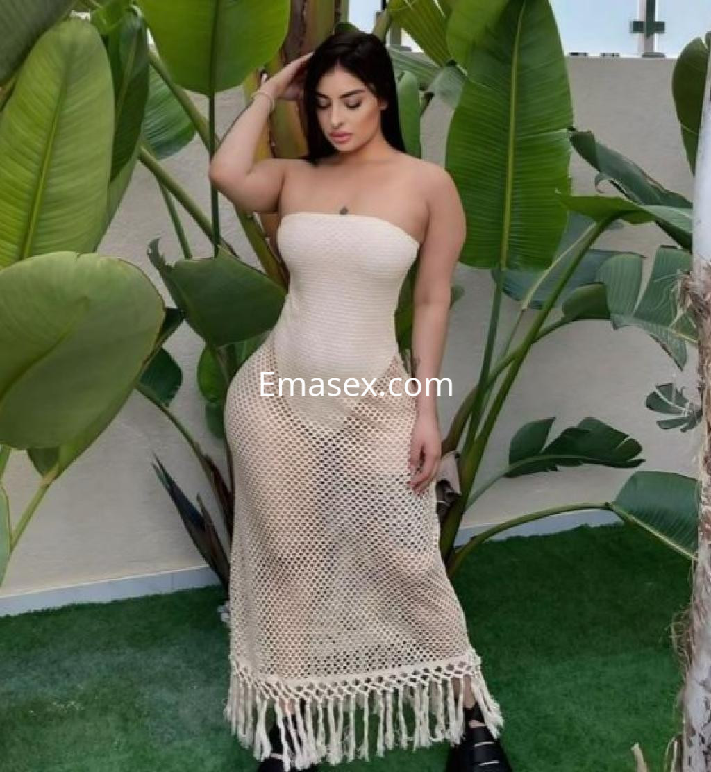 Escort en Alicante posando con un vestido largo de crochet, rodeada de plantas verdes, con cabello oscuro.