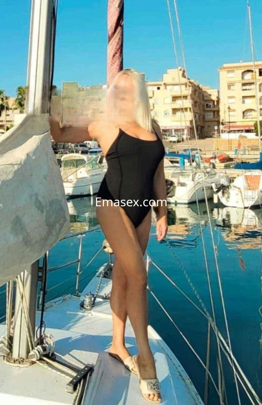 Puta en Almería, posando en un barco con un bañador negro, luciendo un cabello rubio y un entorno marítimo.