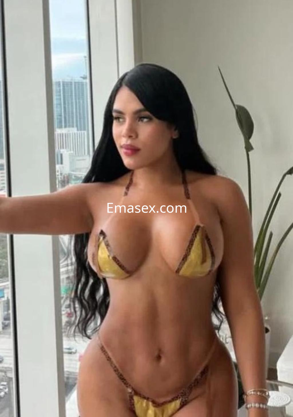 Escort con cabello largo y negro, vistiendo un bikini dorado, posando junto a una ventana en Almería.