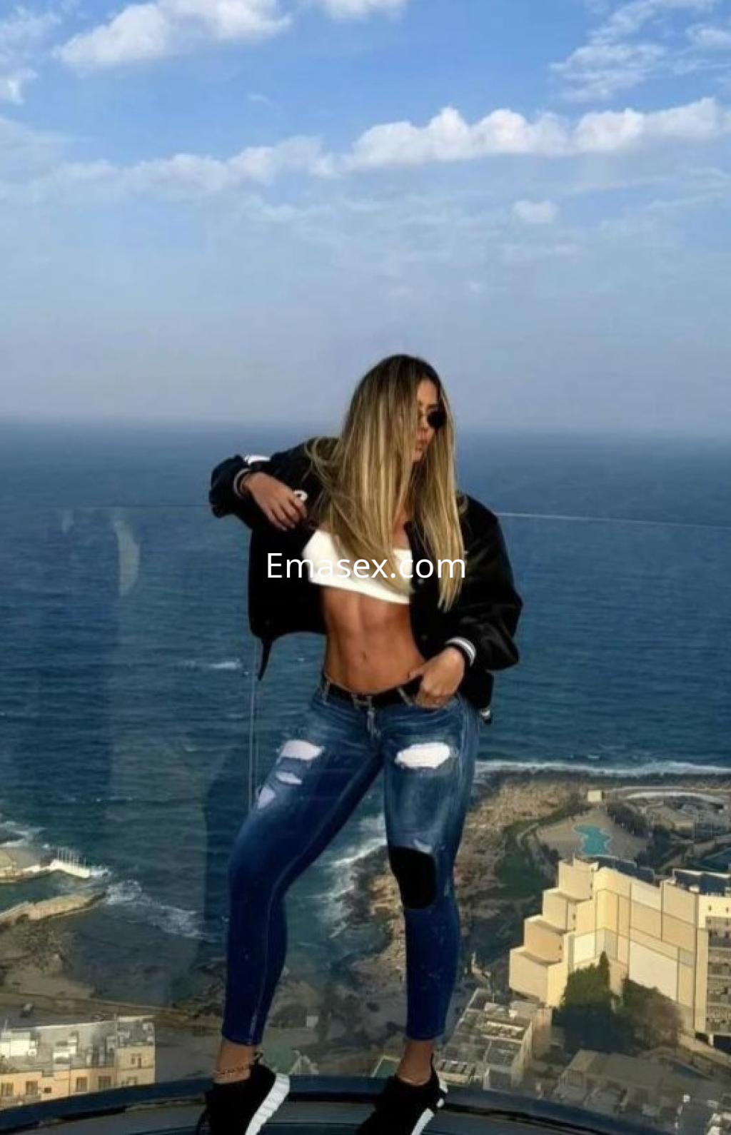 Escort en Tarragona con cabello largo y lacio, vistiendo top blanco y jeans rasgados, posando frente al mar.