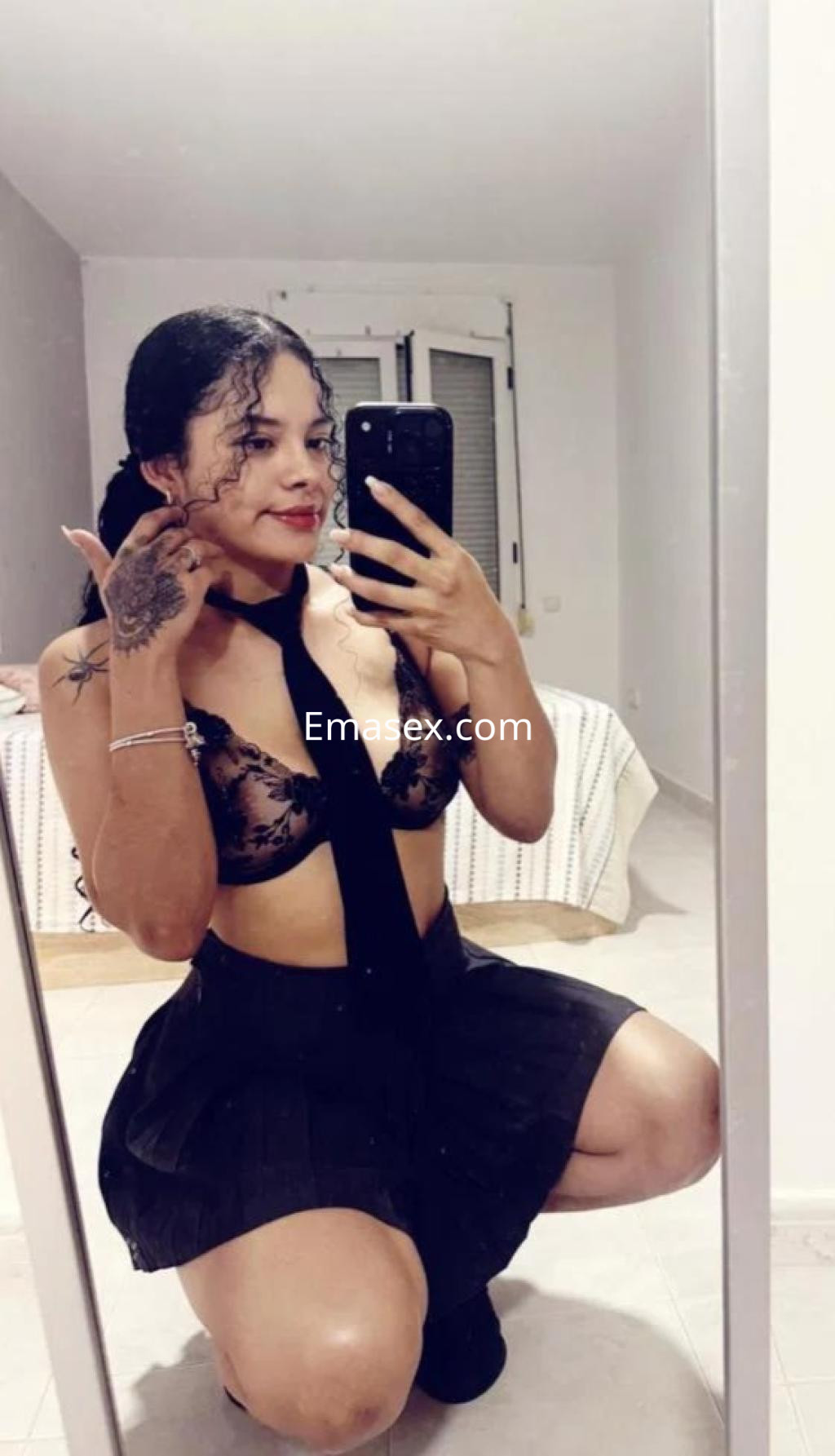 La puta en Tarragona lleva un sujetador negro de encaje y una mini falda, posando con confianza frente al espejo.