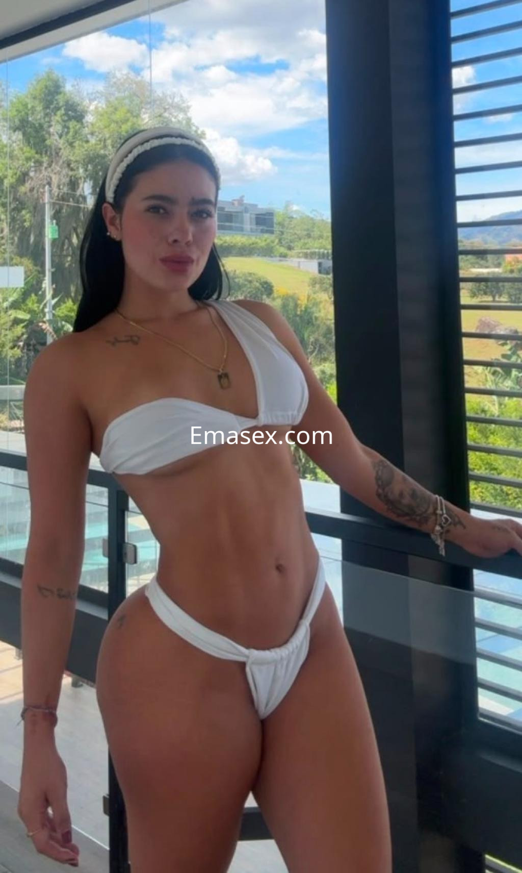 Escort en Barcelona con cabello oscuro y piel bronceada, luciendo un bikini blanco mientras posa en un balcón.