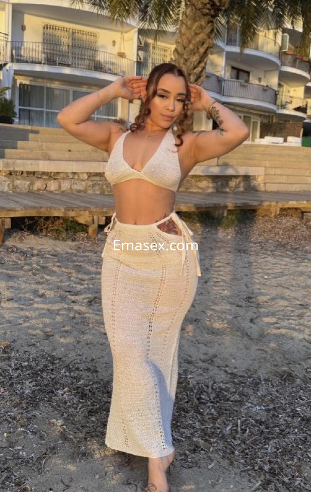 Puta en Barcelona luciendo un conjunto de bikini blanco con falda larga de crochet, posando en la playa.