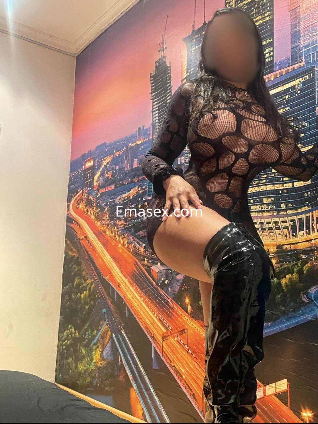 Escort en Zaragoza, vistiendo un vestido de malla negra con patrones circulares y botas altas brillantes, posando de lado.