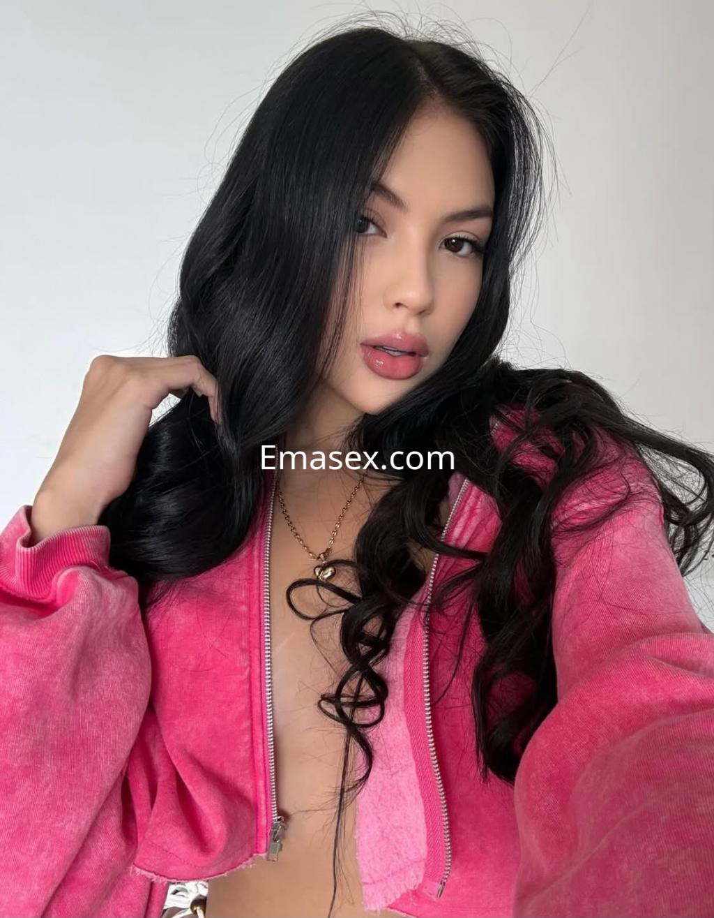 Escort en Pamplona con cabello largo y ondulado, vistiendo una sudadera rosa y posando en un fondo claro.