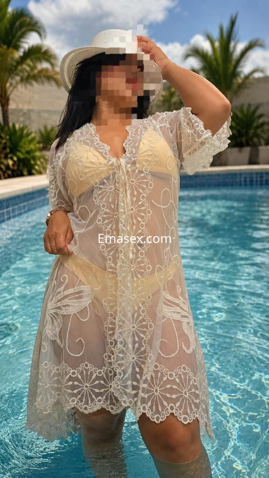 Escort en Pamplona, posando en una piscina con un vestido transparente y sombrero, rodeada de palmeras.