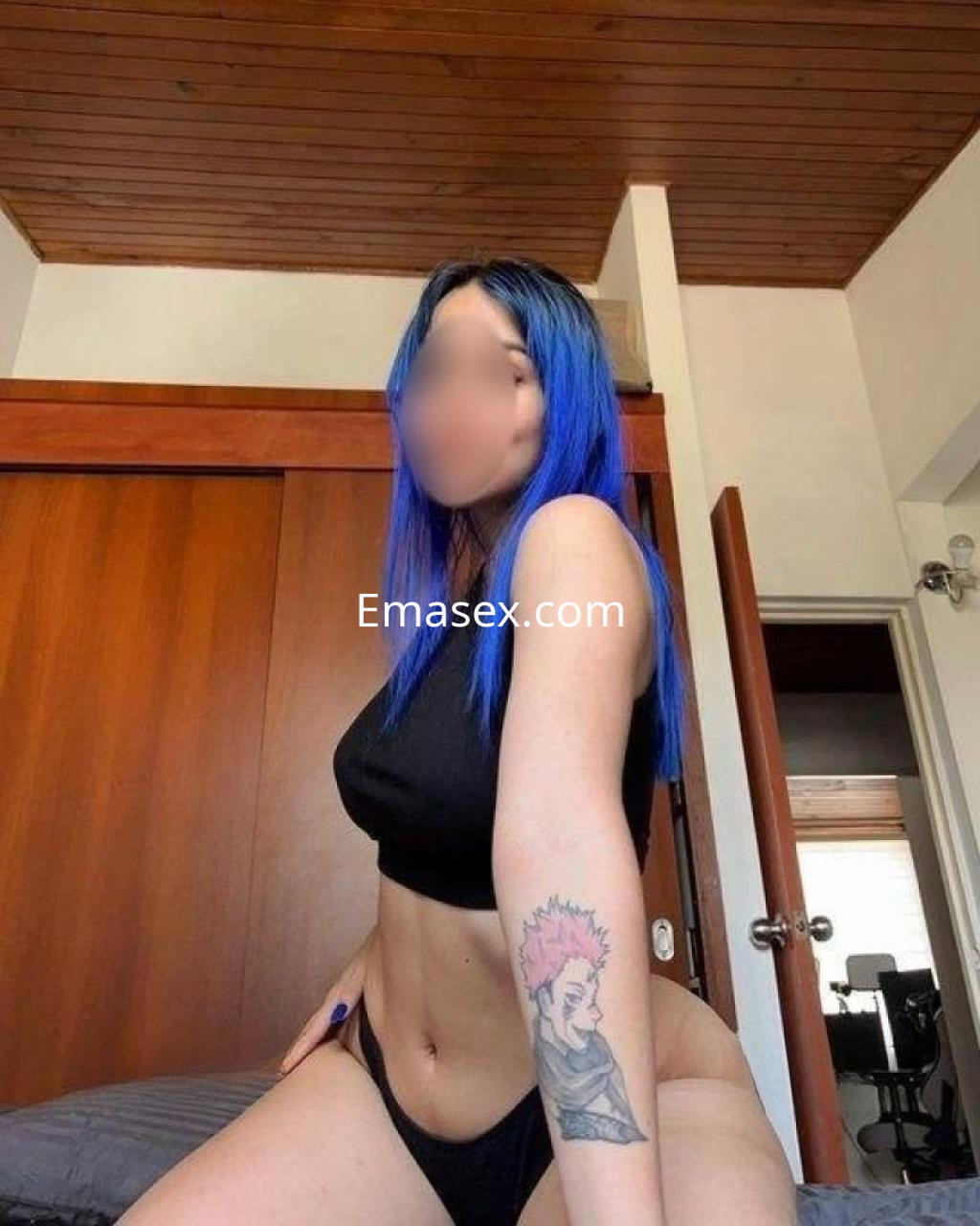 Escort de Las Palmas con cabello azul y tatuaje, vestida con top negro y lencería negra en un entorno interior.