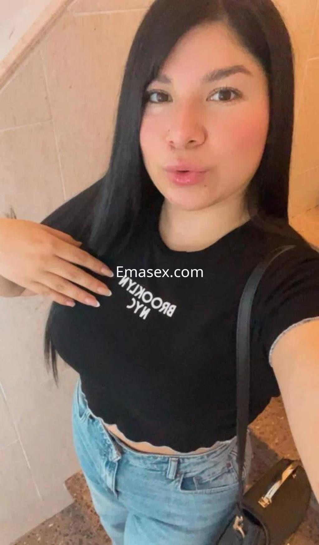 Puta en Barcelona viste camiseta negra con texto y jeans claros, posando con una mano en el pecho.