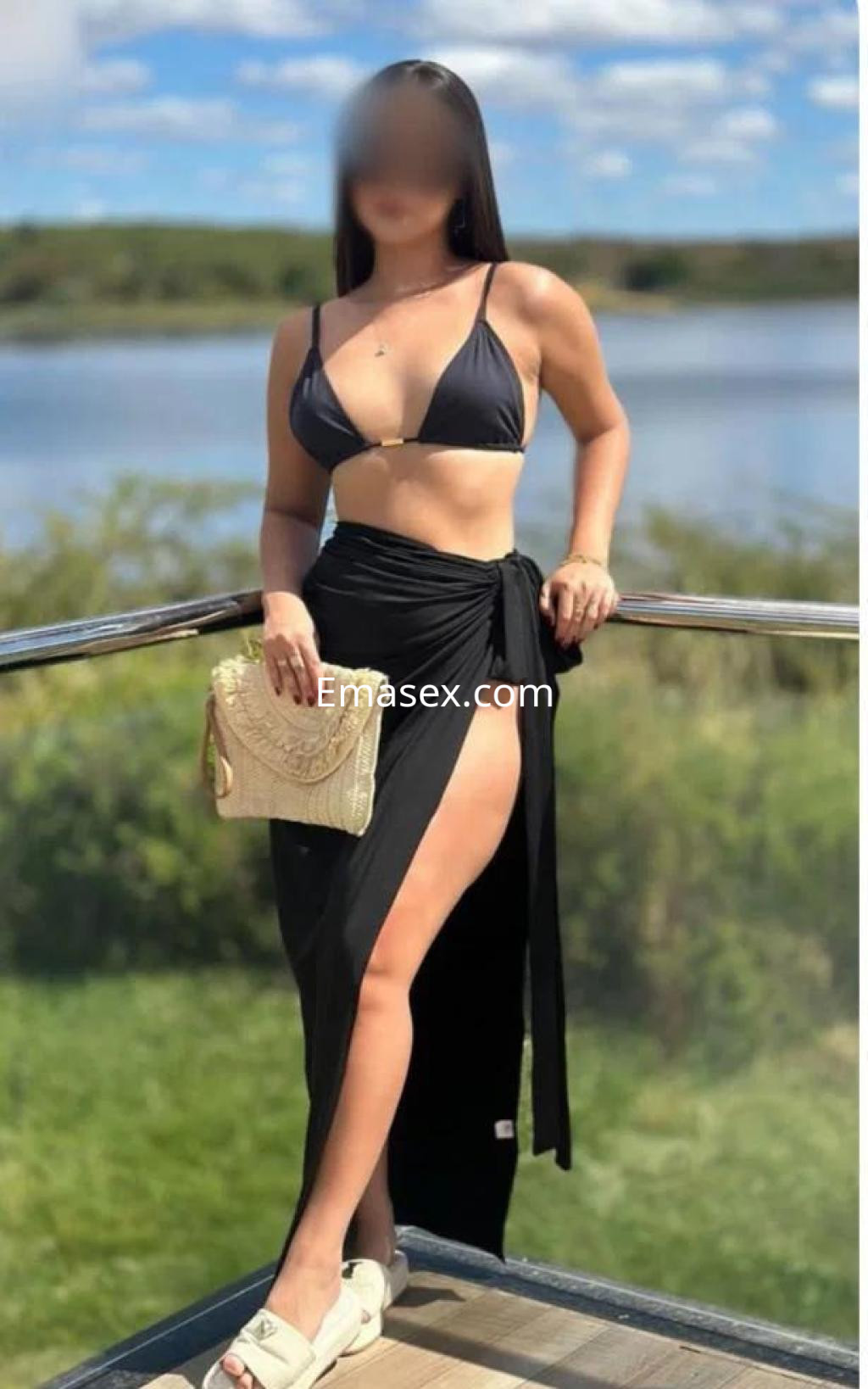 Escort en Girona con top negro y falda larga, posando de pie junto a un lago, con un bolso claro en la mano.