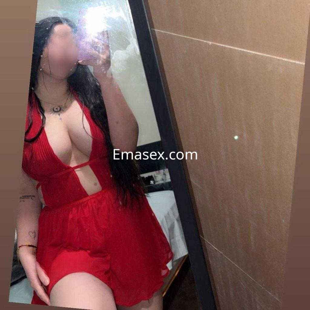 Escort en León posando frente al espejo con un vestido rojo que resalta su figura y cabello largo.