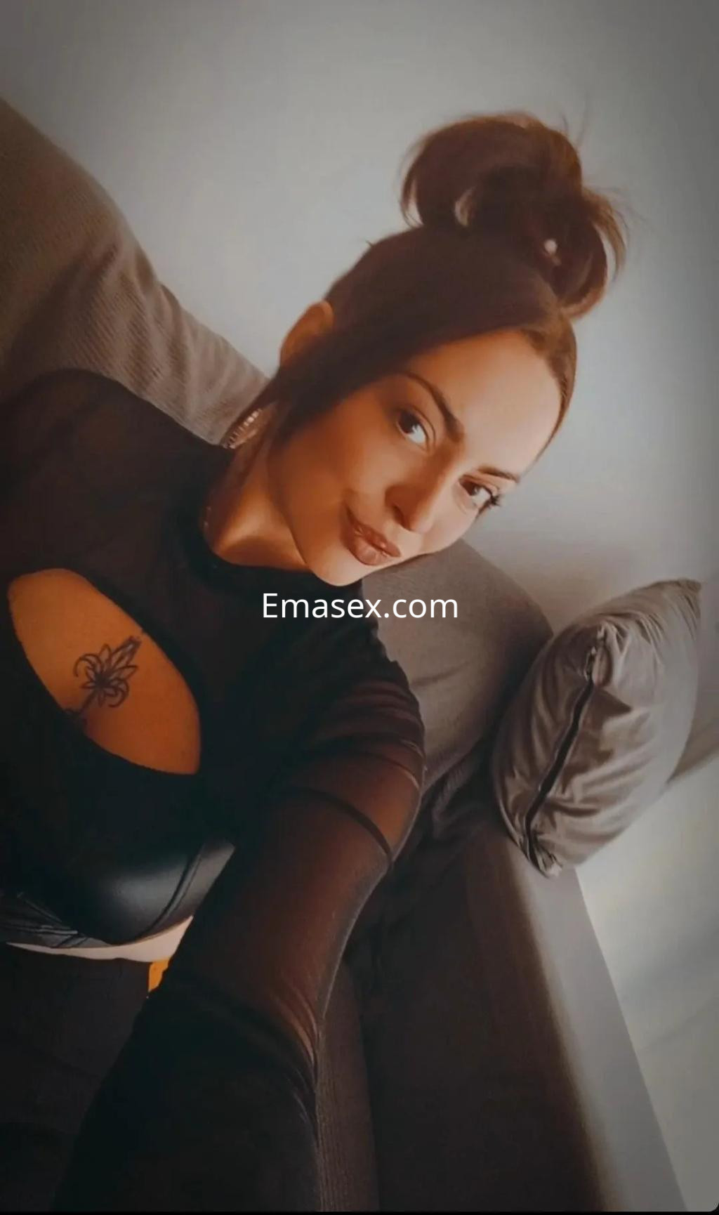 Escort en Sevilla con blusa negra transparente y peinado recogido, sonriendo en un sofá.