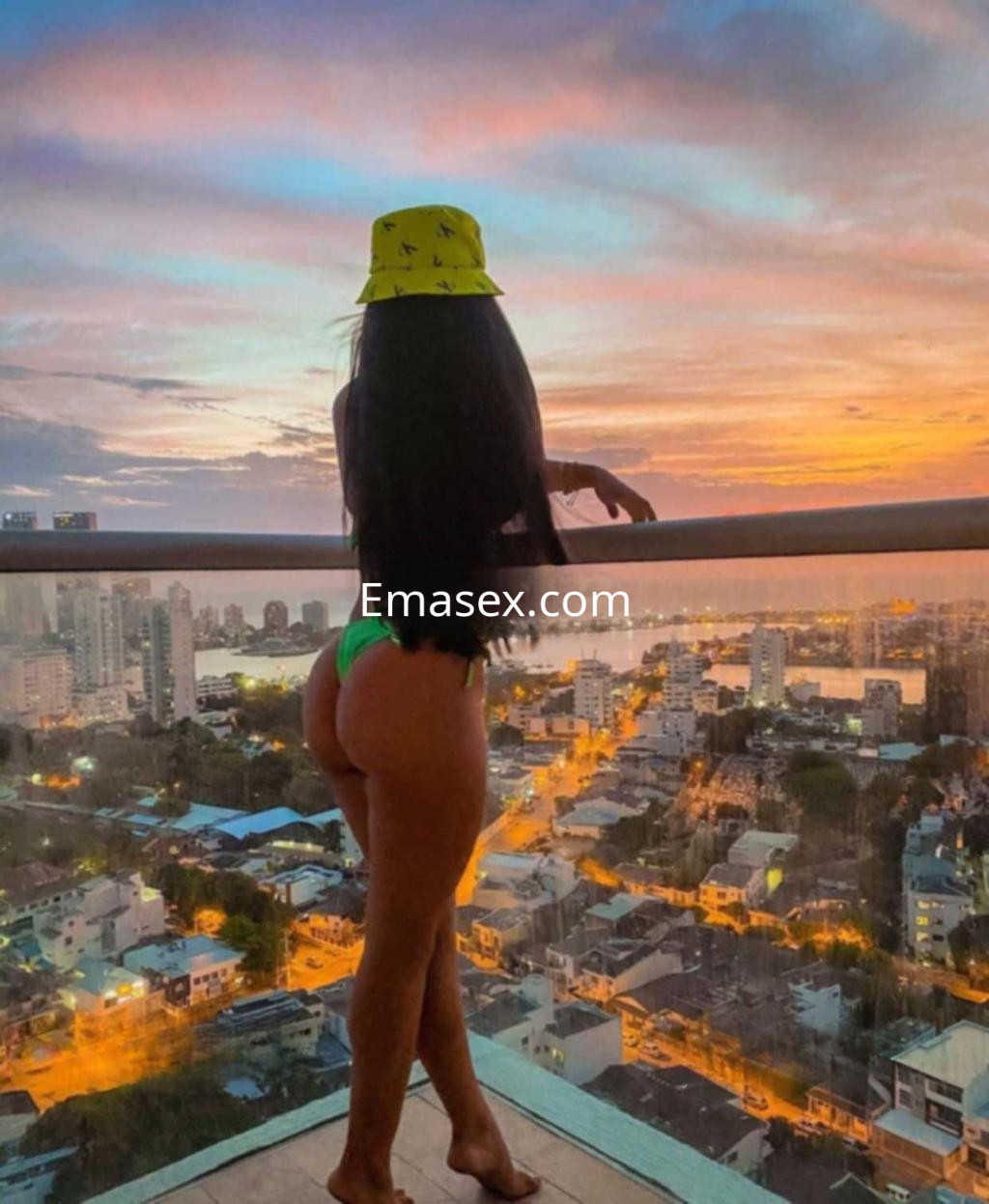 Puta en Toledo, de espaldas con un sombrero amarillo y bikini verde, contemplando un atardecer urbano.