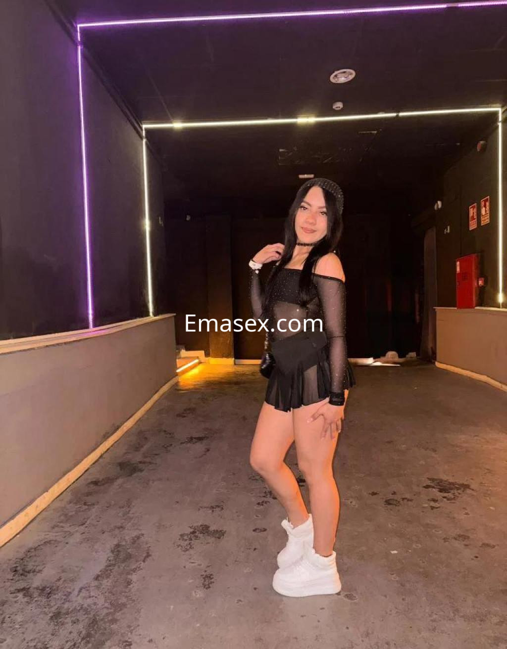 Puta en Valencia con cabello largo y lacio, luciendo un atuendo negro y zapatillas blancas en un entorno iluminado.