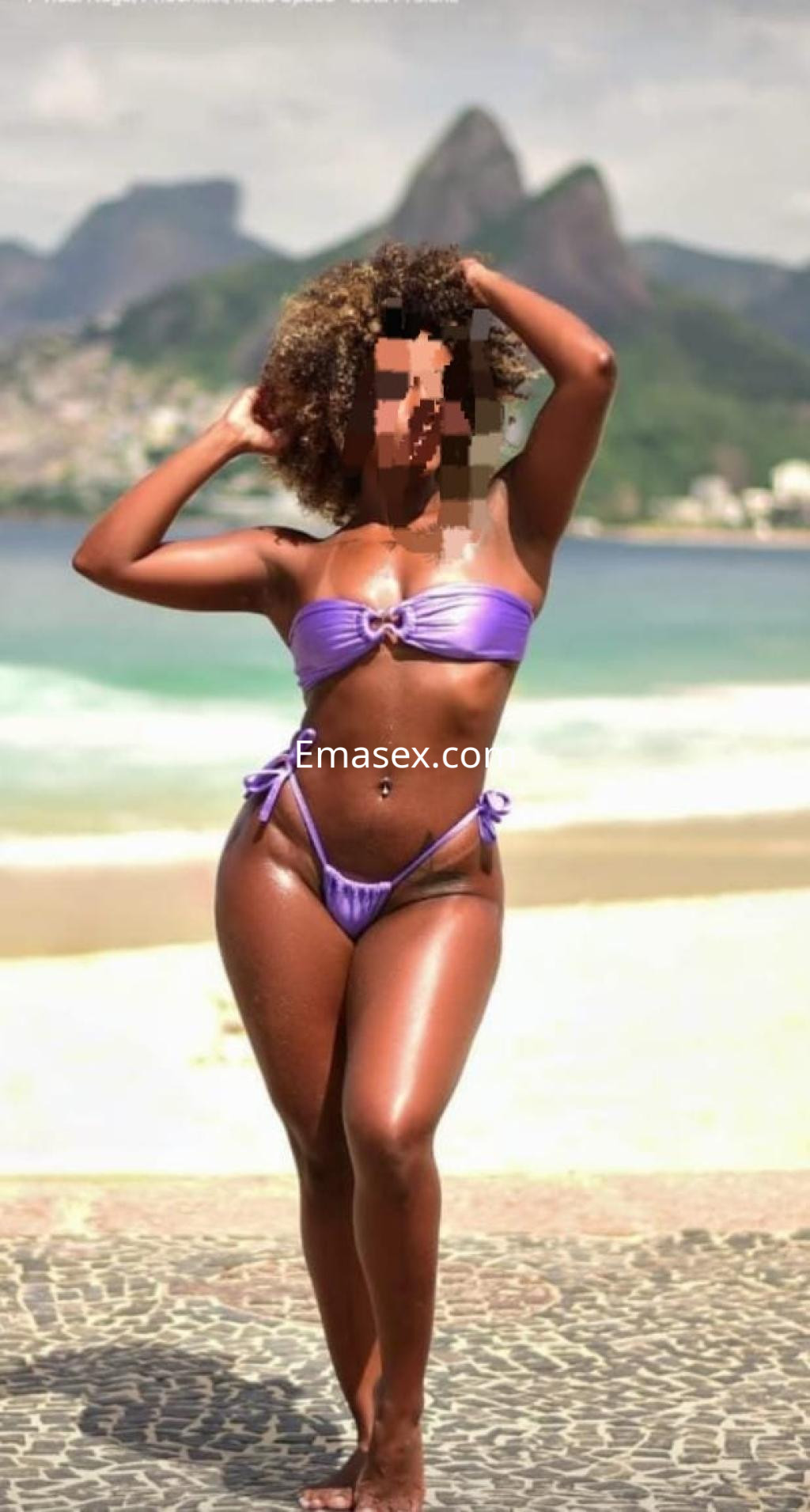 Puta en Coruña luciendo un bikini morado, posando con confianza en la playa y un paisaje montañoso al fondo.