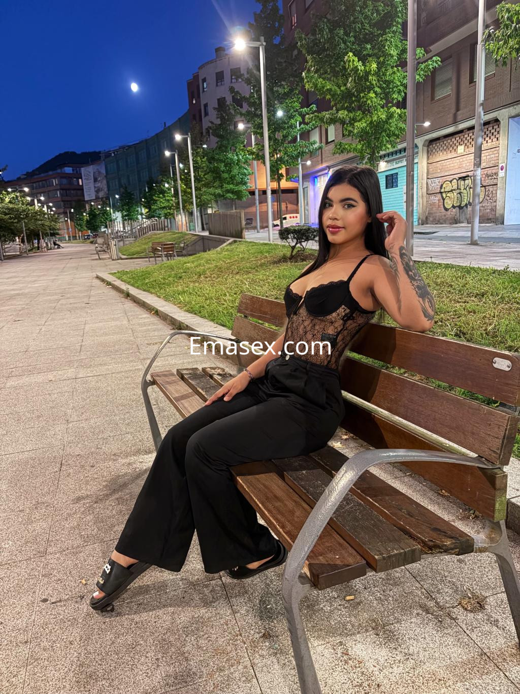 Puta en Bilbao vestida con un body negro de encaje y pantalones negros, sentada en un banco al aire libre.
