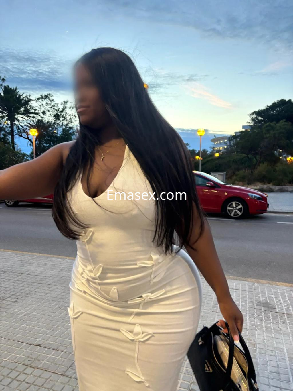 Escort de Palma de Mallorca posando con un vestido blanco ceñido y llevando un bolso negro, con un fondo urbano.