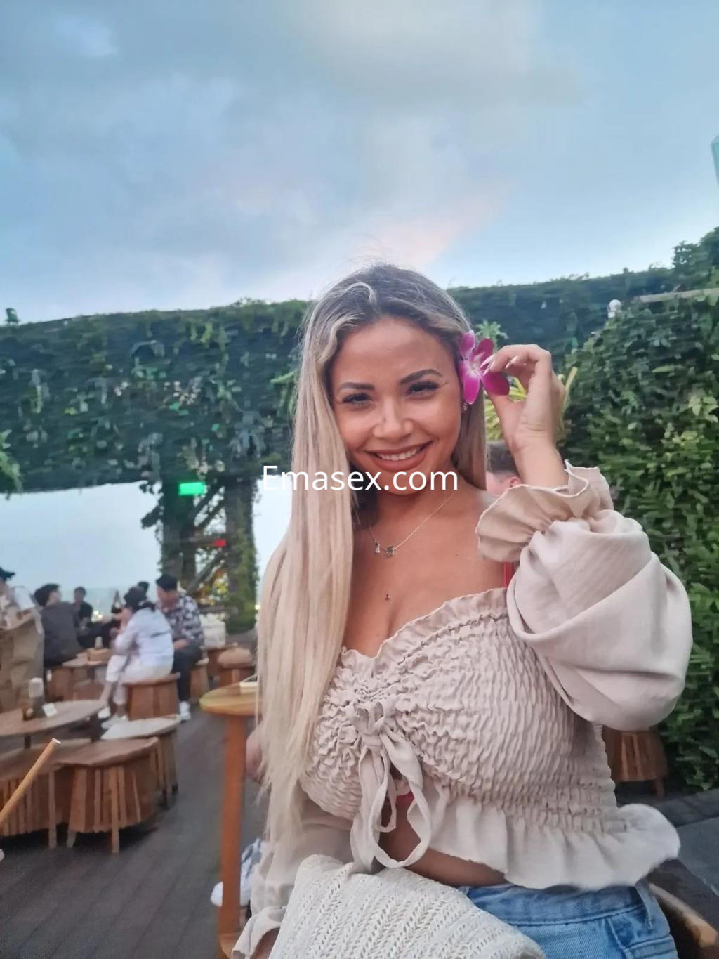 Escort en Palma de Mallorca con blusa clara y flor en el pelo, sonriendo en un ambiente al aire libre.