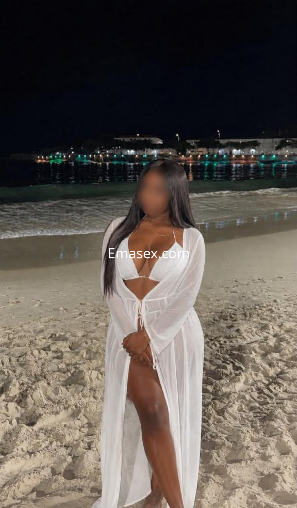 Puta de pie en la playa de Tenerife, con un vestido blanco y cabello largo, posando junto al mar de noche.