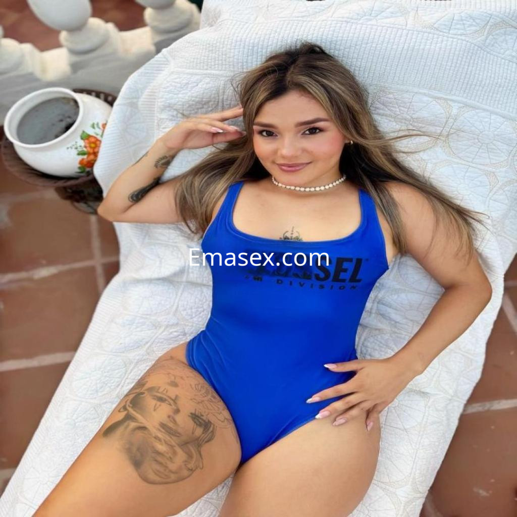 Escort en Tenerife, posando sobre una tumbona con un traje de baño azul, cabello largo y tatuajes visibles.