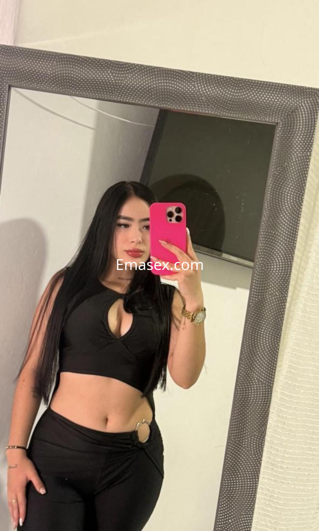 Escort en Tenerife posando frente a un espejo, lleva un top negro con escote y pantalones ajustados, cabello largo y lacio.