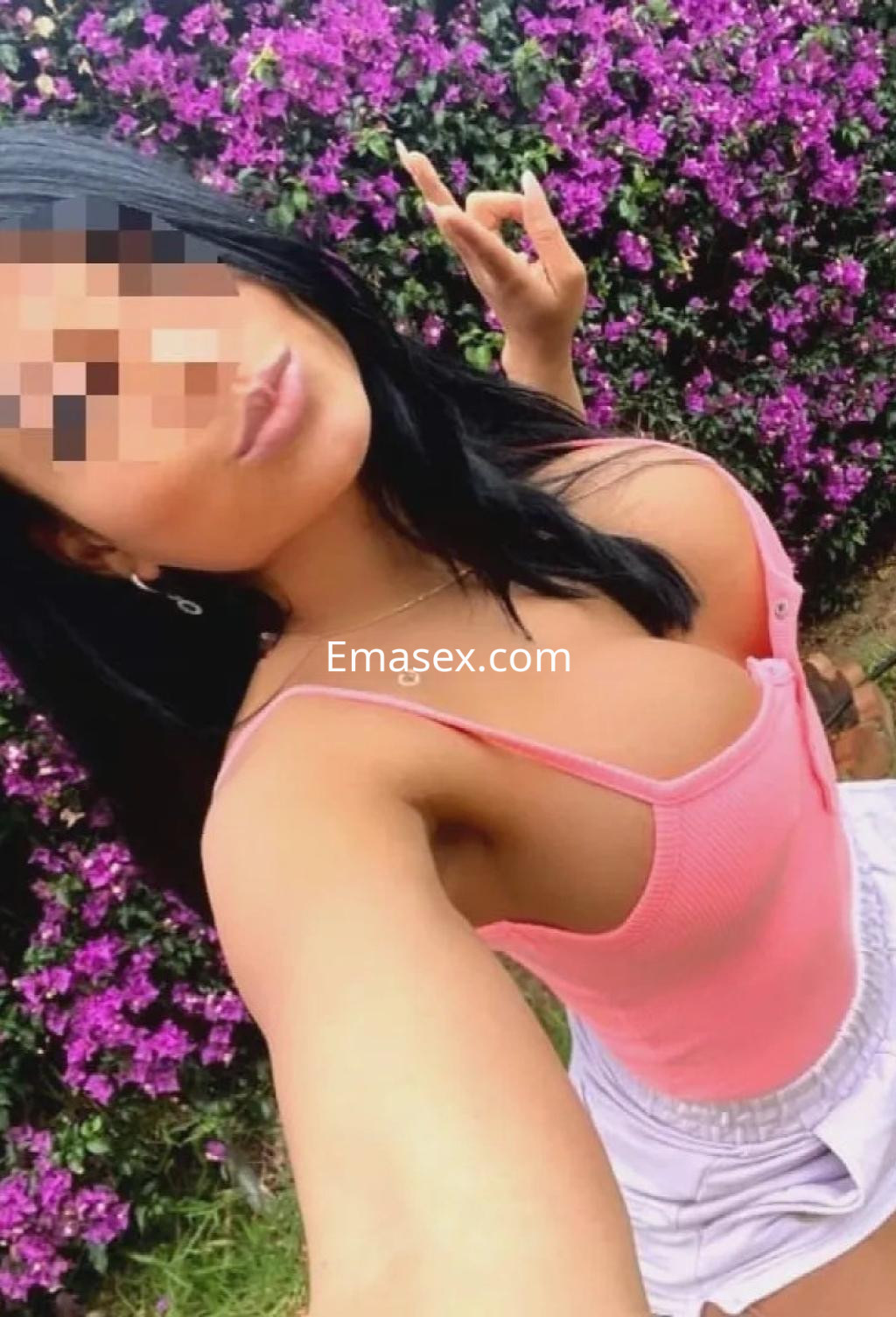 Escort en Málaga con top rosa y falda blanca, posando frente a un fondo de flores moradas.