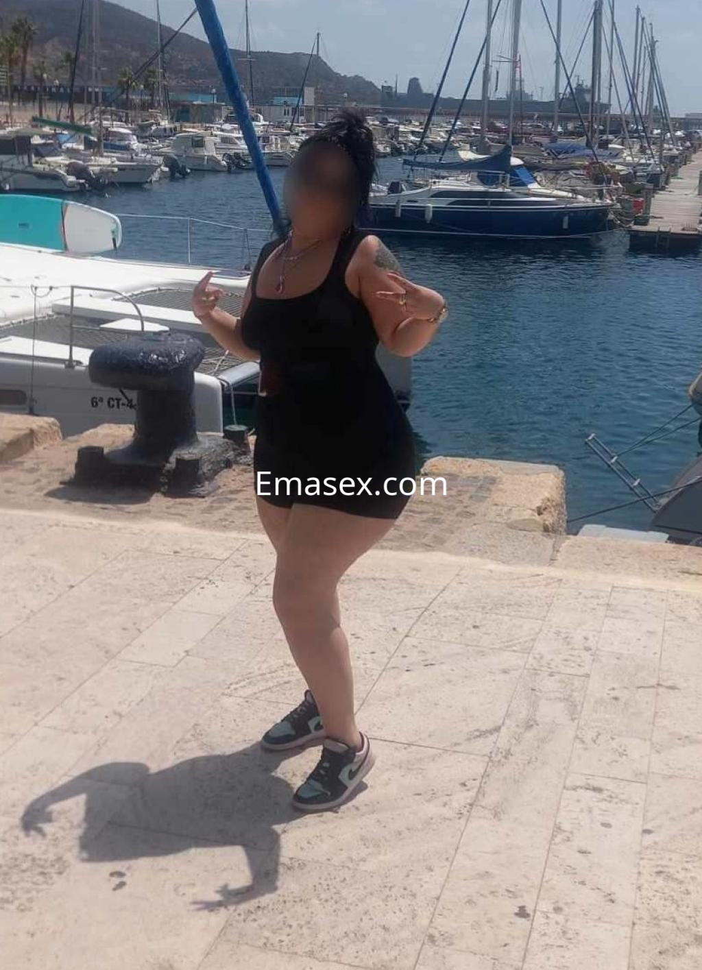 Puta en Málaga, luciendo un vestido negro ajustado y zapatillas deportivas, posando en el puerto con barcos de fondo.