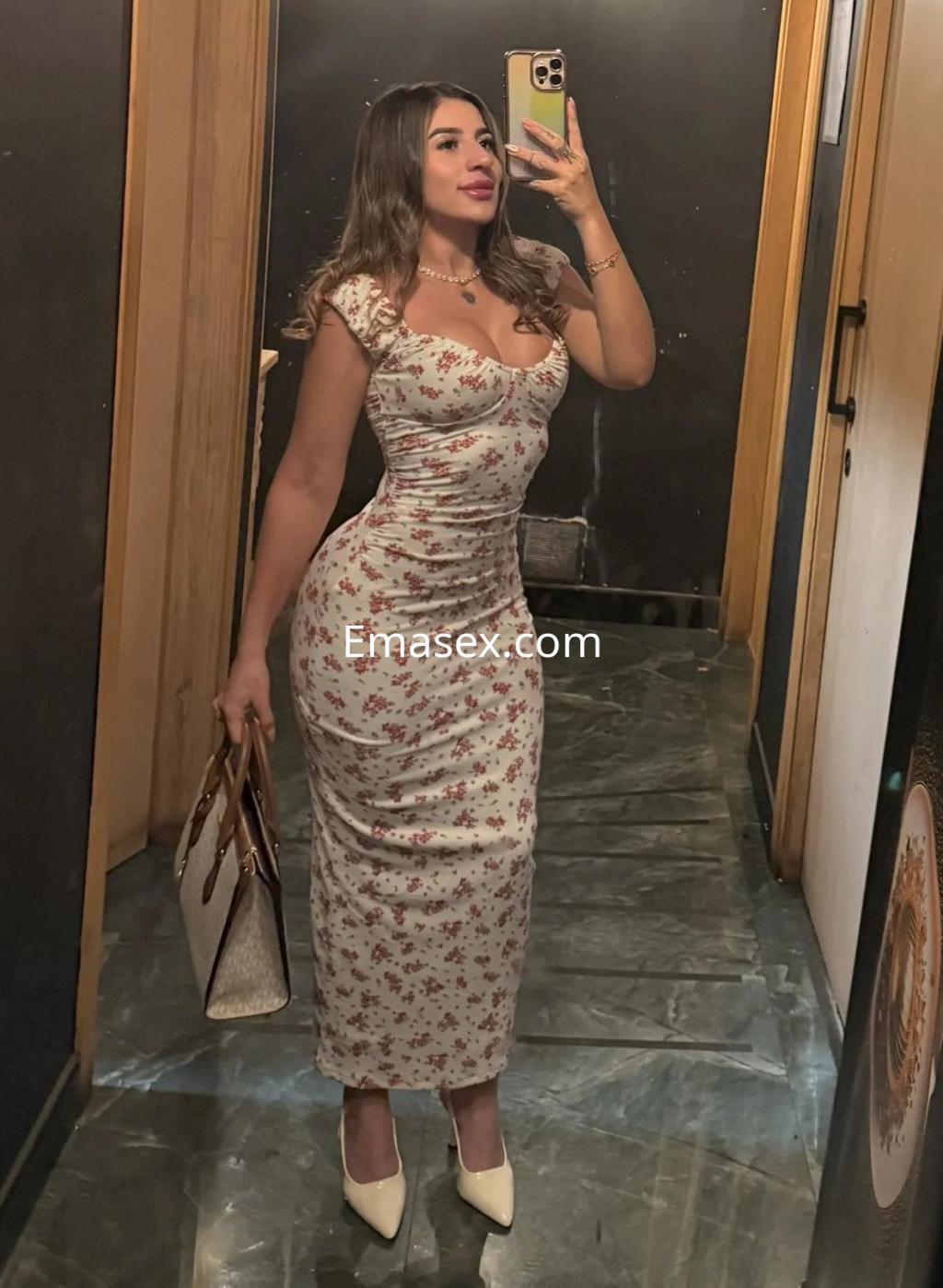 La puta en Málaga luce un vestido ajustado con estampado floral y tacones mientras se toma una selfie en un espejo.