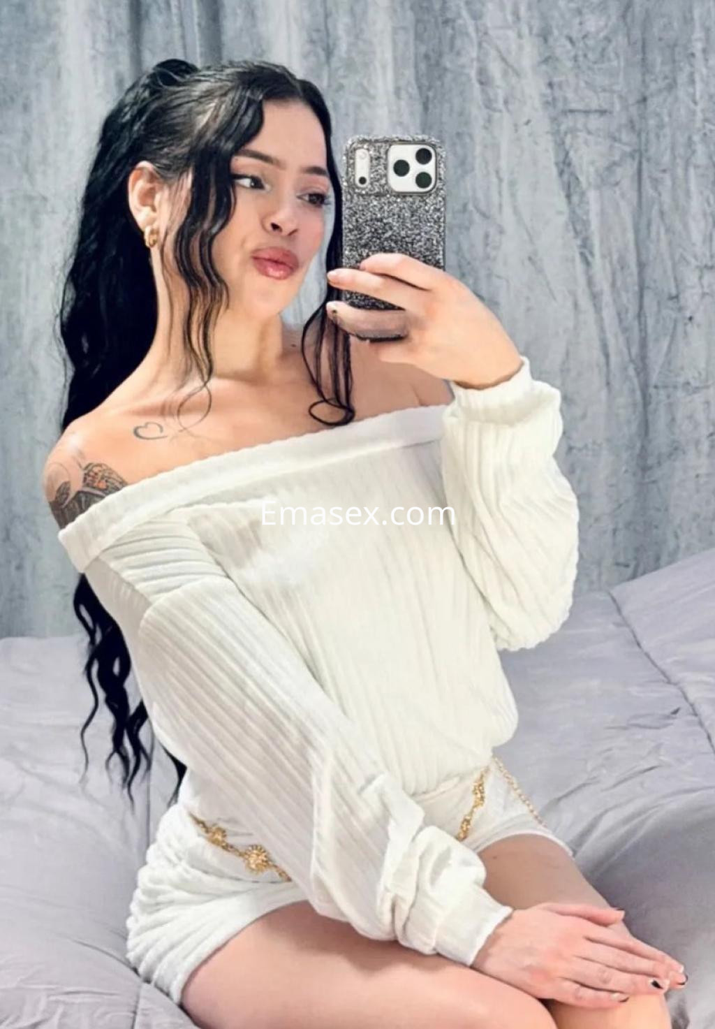 Escort en Málaga con cabello largo y rizado, viste una blusa blanca y shorts, posando frente a un espejo.
