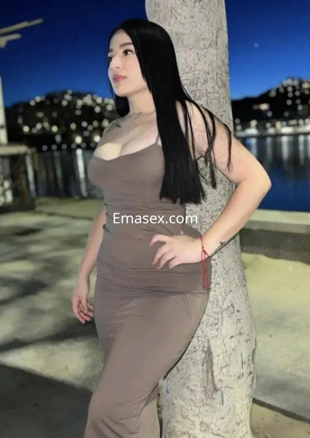La puta se apoya en un árbol con un vestido ajustado color beige, frente a un paisaje nocturno en Oviedo.