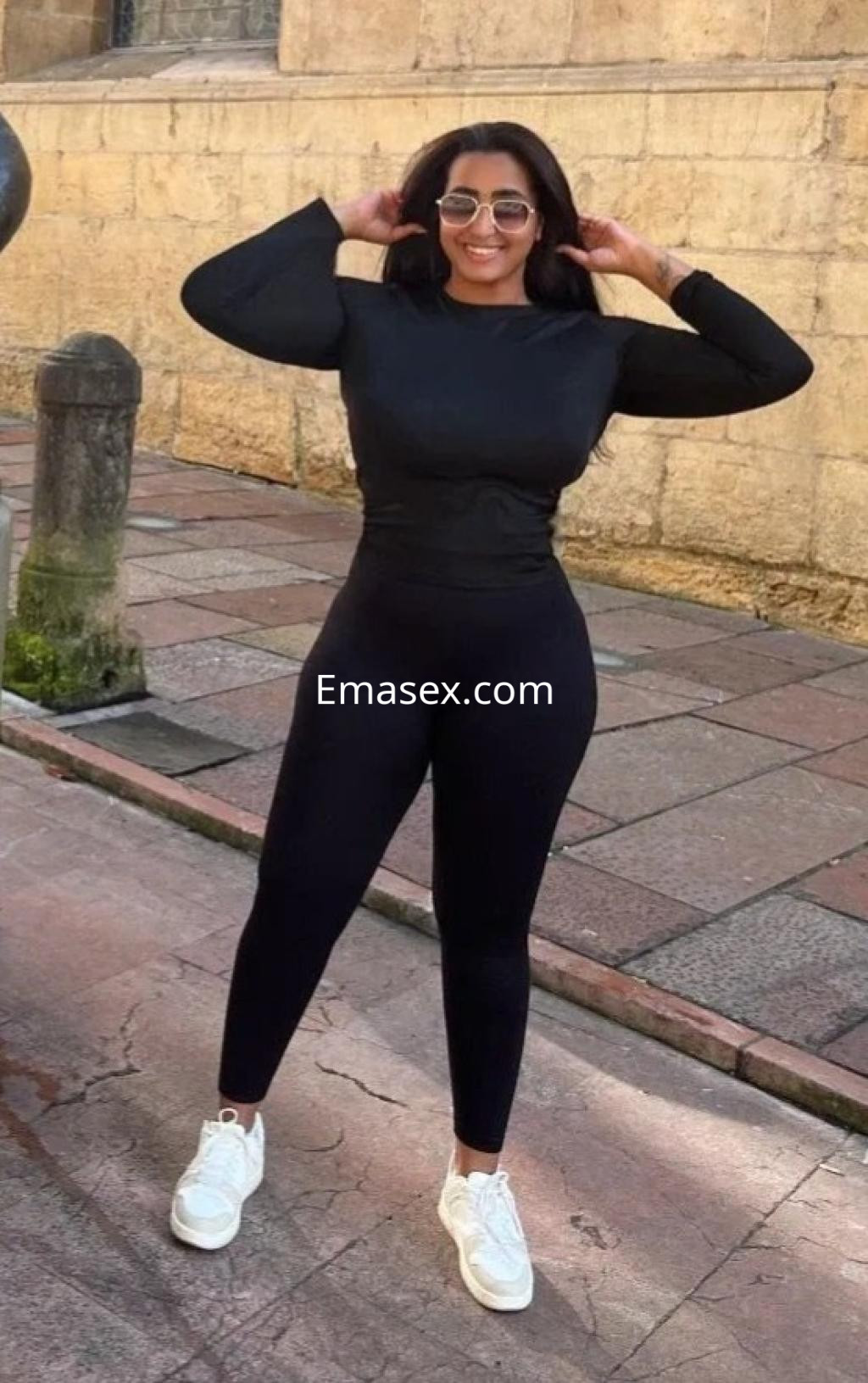 Escort en Oviedo, posando con una camiseta de manga larga y pantalones ajustados negros, con zapatillas blancas.