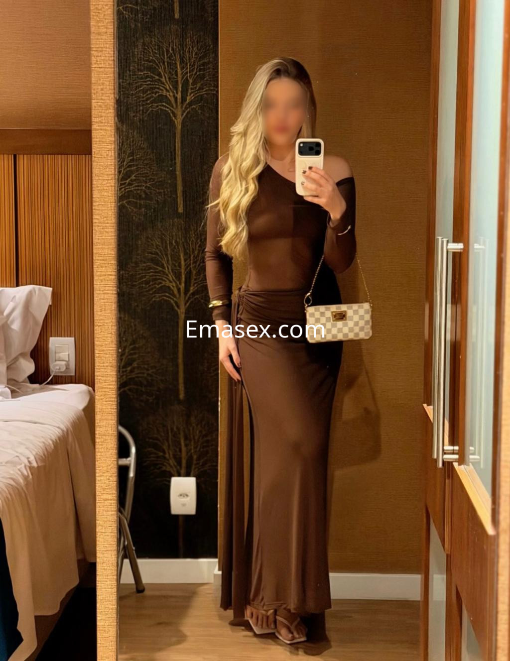 Escort en Donosti, con cabello largo y rubio, viste un vestido largo marrón y sostiene un bolso claro, posando en un hotel.