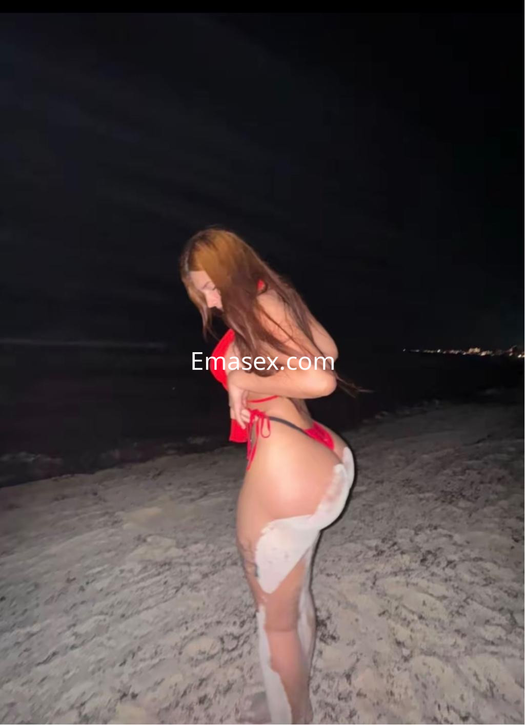 La puta en Pamplona se muestra de lado en una bikini roja en la playa, destacando su figura bajo la luz nocturna.
