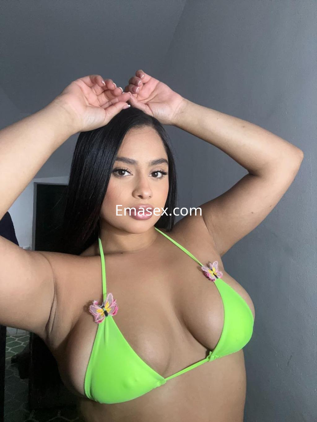 Escort con cabello largo y lacio, viste un bikini verde con detalles de mariposas, en un ambiente iluminado de Vitoria.