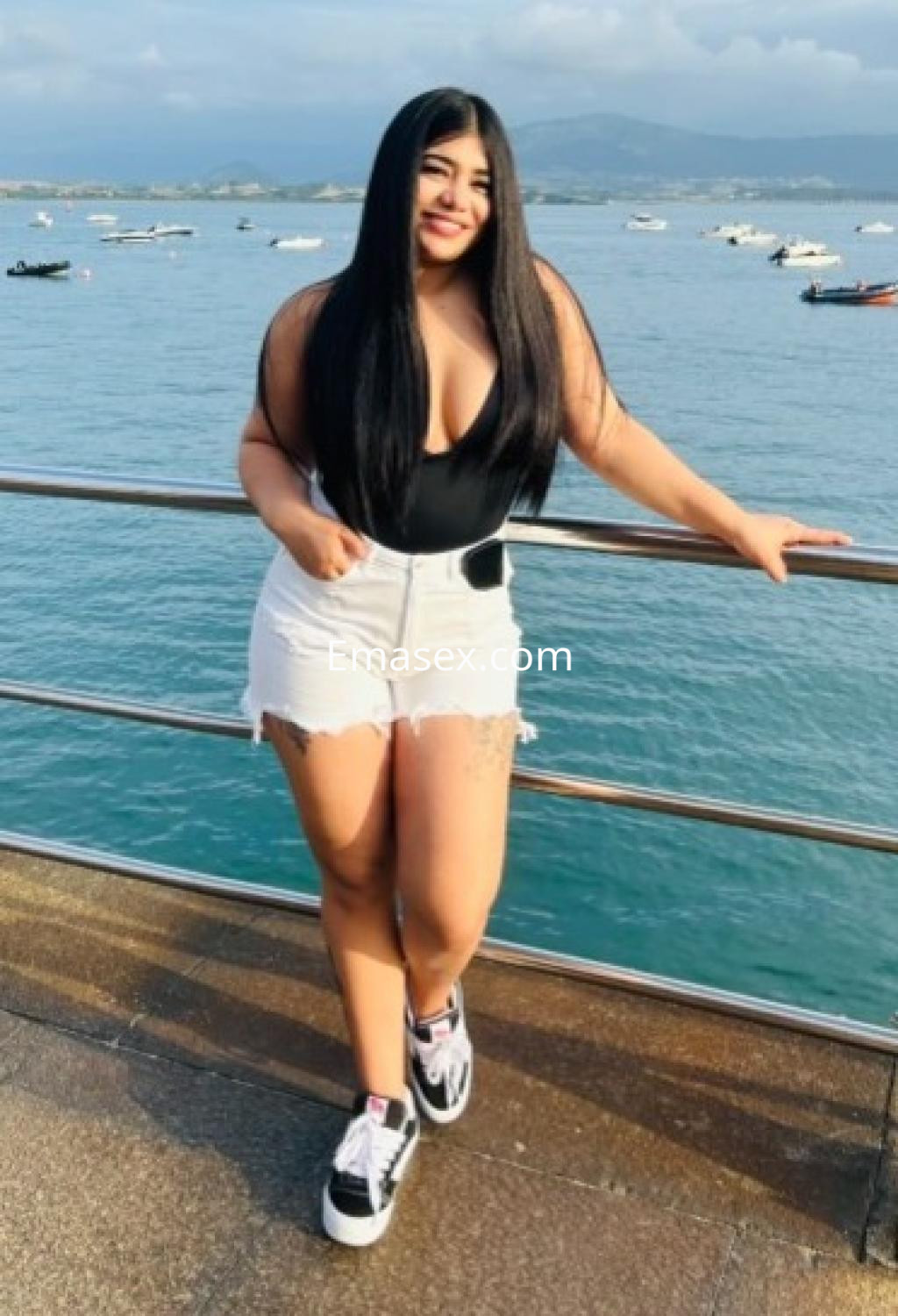 Escort en Vitoria, con cabello largo y oscuro, viste un body negro y shorts blancos, posando junto al mar.