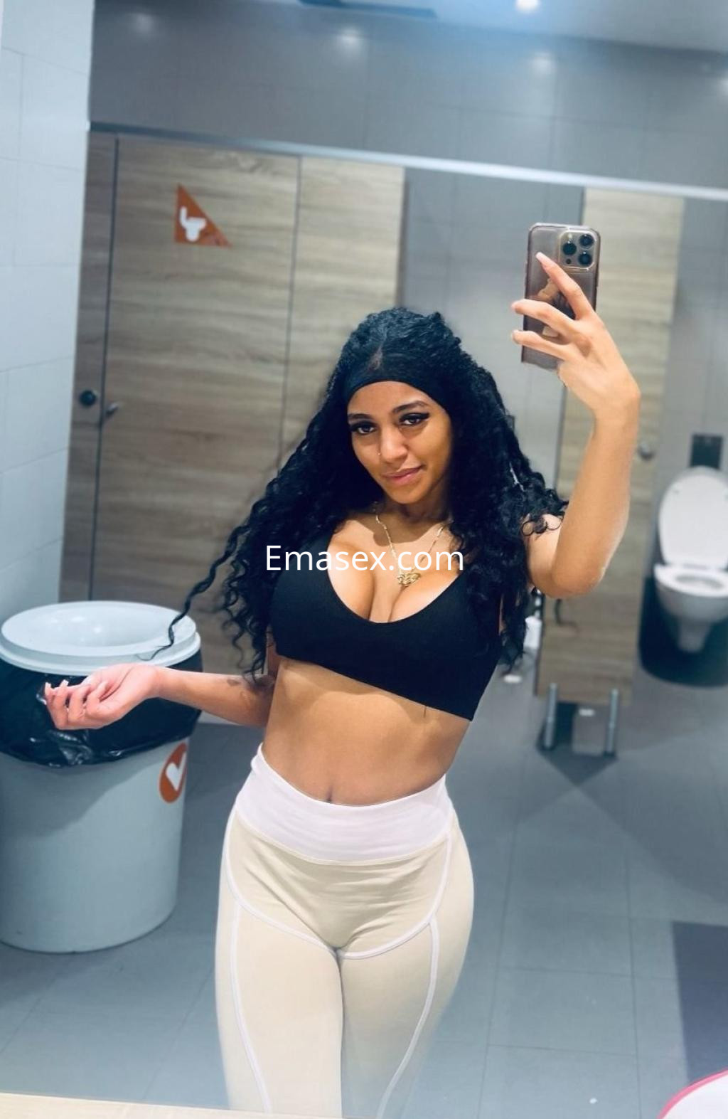 Escort en Palma de Mallorca posando frente al espejo en un baño, con top negro y leggins claros, sonriendo.