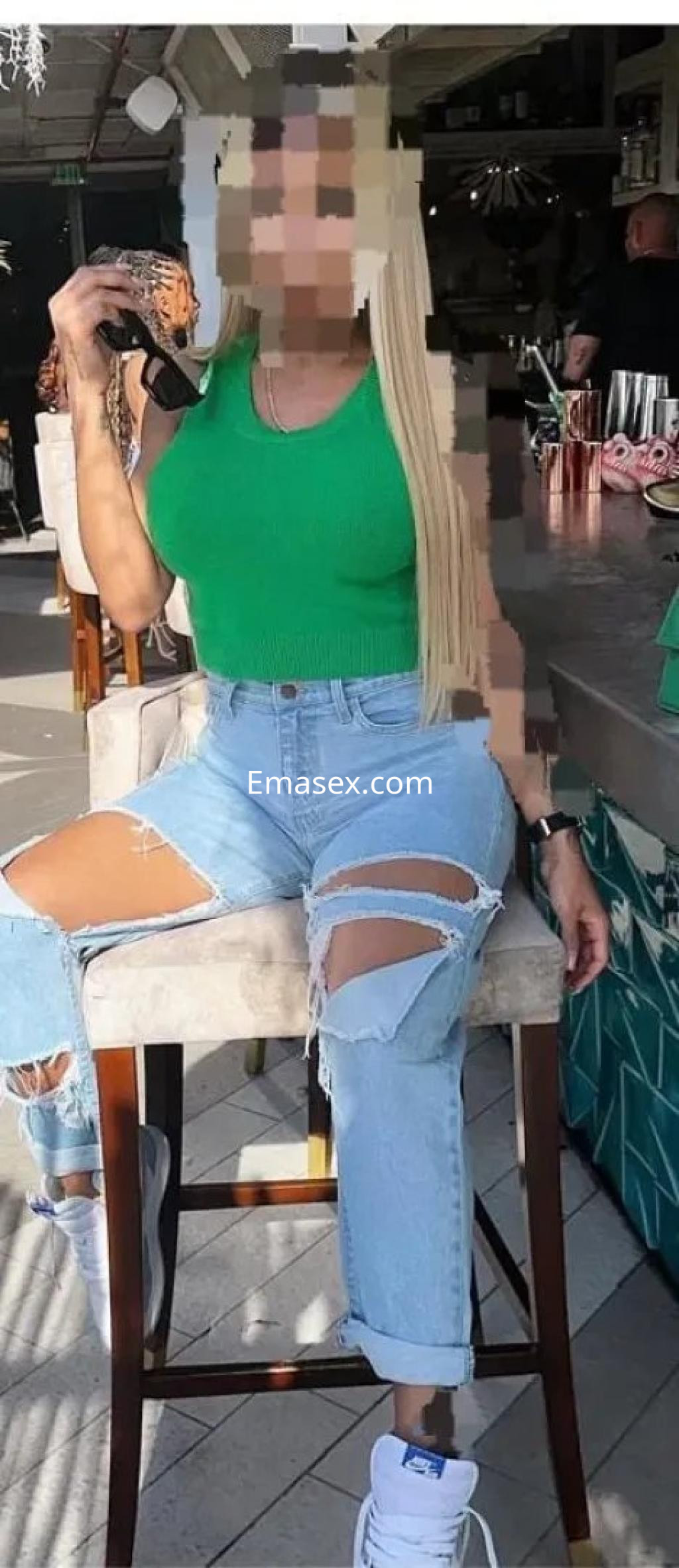 Puta en Albacete, sentada en una barra, viste un top verde y jeans rasgados, luciendo cabello rubio largo.