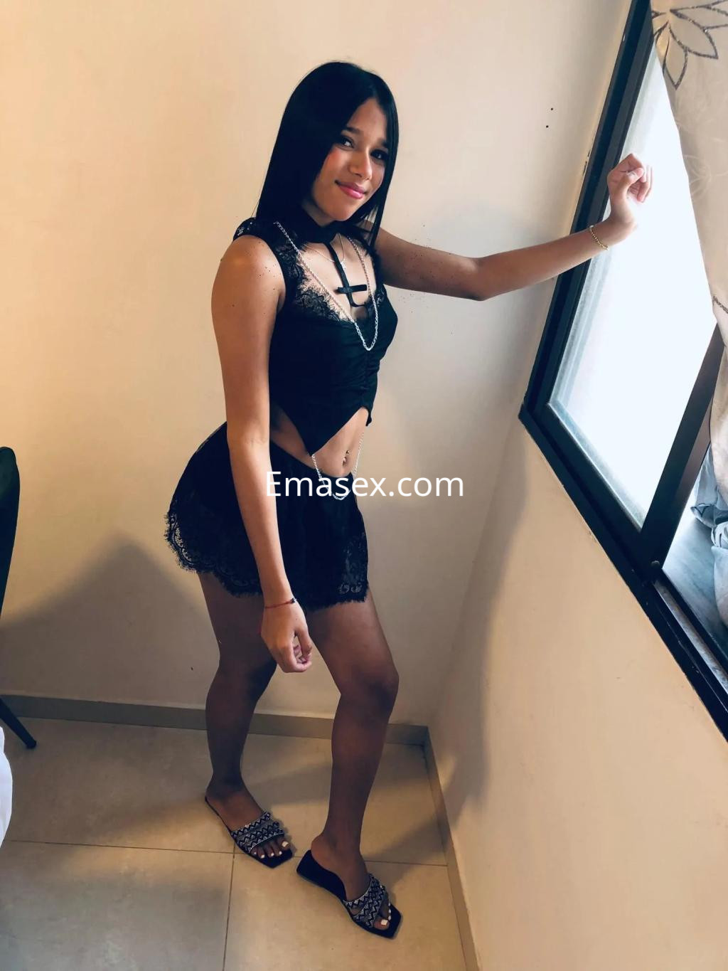 Puta posando junto a una ventana en Alicante, vistiendo un conjunto de lencería negra y sandalias.
