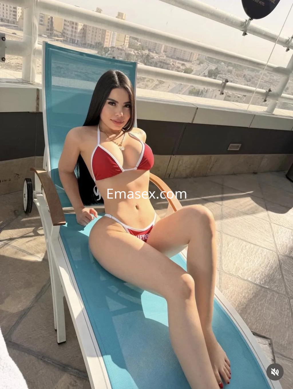 Escort en Alicante luciendo un bikini rojo y blanco, relajada en una tumbona junto a una vista urbana.