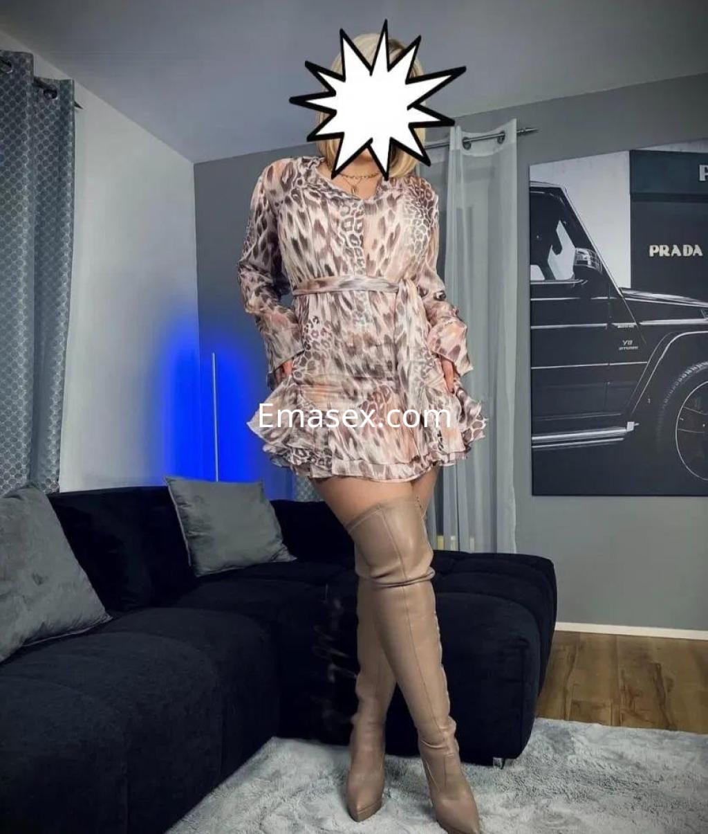 Puta en Badajoz posando con un vestido claro y botas altas, en un ambiente moderno con sofá oscuro.