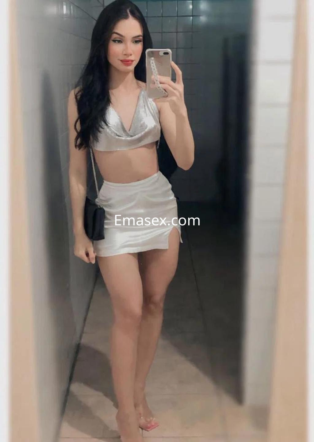 Puta en Barcelona con top plateado y falda corta, posando en un baño con azulejos claros y un bolso negro.