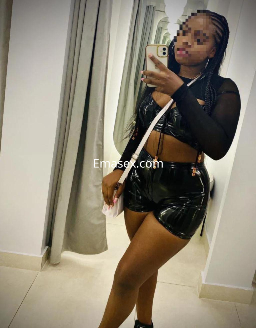 Escort en Girona posando en un espejo con un conjunto de lencería negra brillante y sandalias.