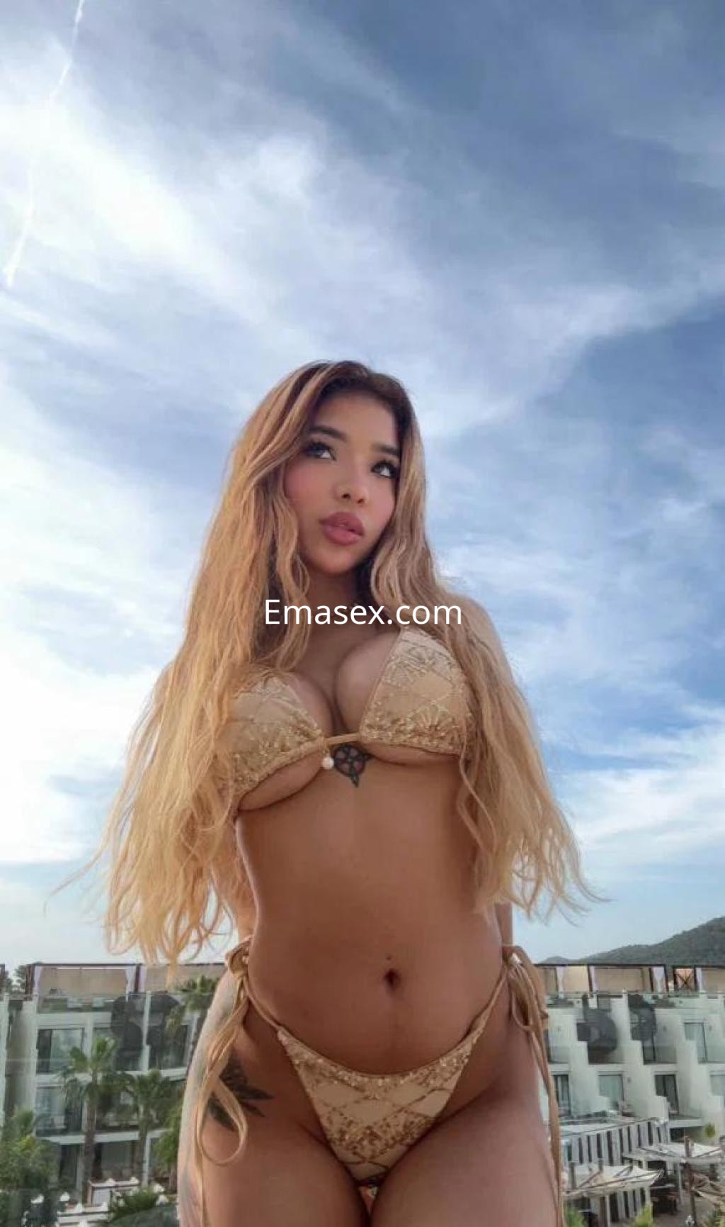La puta luce un bikini dorado, con cabello largo y suelto, posando frente a un cielo despejado en Granada.