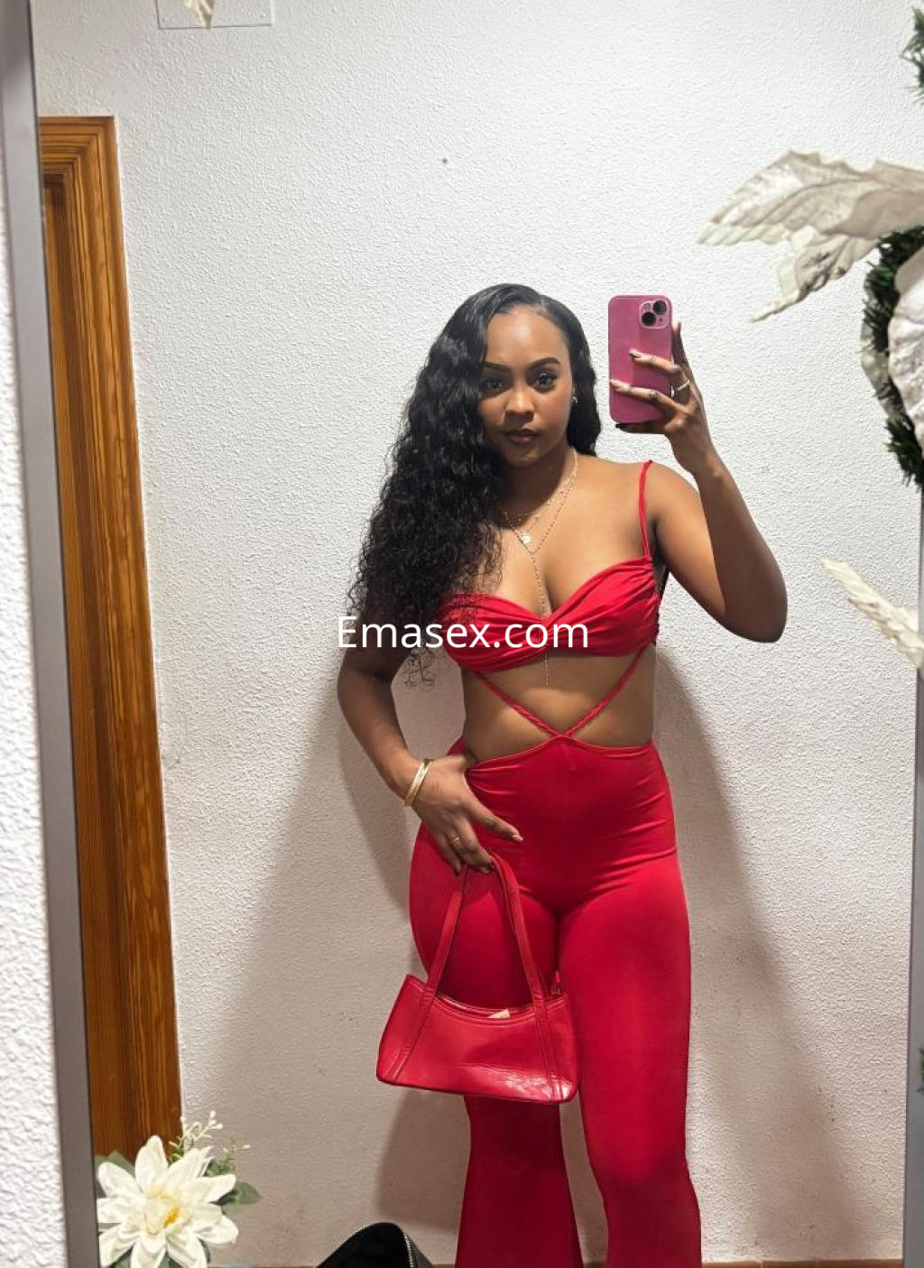 Escort en Lleida posando en un espejo con un conjunto rojo ceñido y un bolso a juego, luciendo pelo rizado.